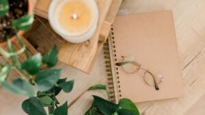 Brennende Kerze und Notizbuch mit einer Brille darauf. Alles ist in beige gehalten